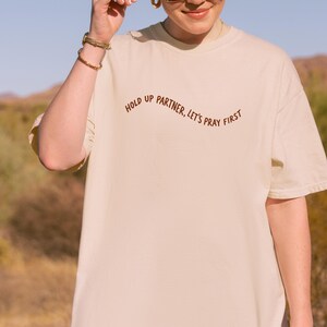 May include: A beige t-shirt with the text "HOLD UP PARTNER, LET'S PRAY FIRST" in brown script. The shirt is made of a soft material and has short sleeves. The image is taken outdoors in natural light.