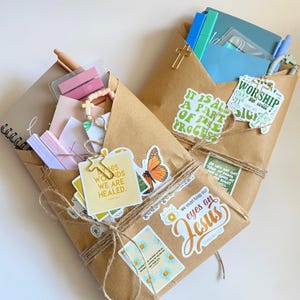 May include: Two brown paper packages tied with twine, containing notebooks, pens, and stationery. The packages are decorated with various stickers featuring religious text, floral designs, and a butterfly, along with small gold cross clips.