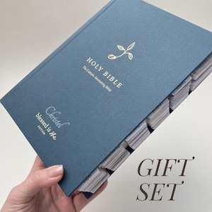 Puede incluir: Una Biblia de tapa dura azul, The Catholic Annotating Bible, con texto en lámina dorada y un pequeño icono de hoja en la portada. El libro incluye el texto Christel Blessed is she EDITION y tiene pestañas de índice blancas a lo largo del borde.