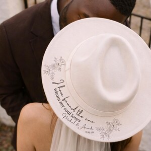 Puede incluir: Un sombrero fedora blanco con detalles florales y el texto "I have found the one whom my soul loves Michael & Amanda 11.15.28". Un accesorio elegante para una boda o evento especial.