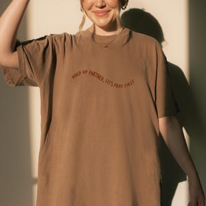 May include: A brown t-shirt with the text "HOLD UP PARTNER, LET'S PRAY FIRST" in a curved, rust-colored font. The shirt has short sleeves and a crew neck. The fabric appears soft and comfortable.