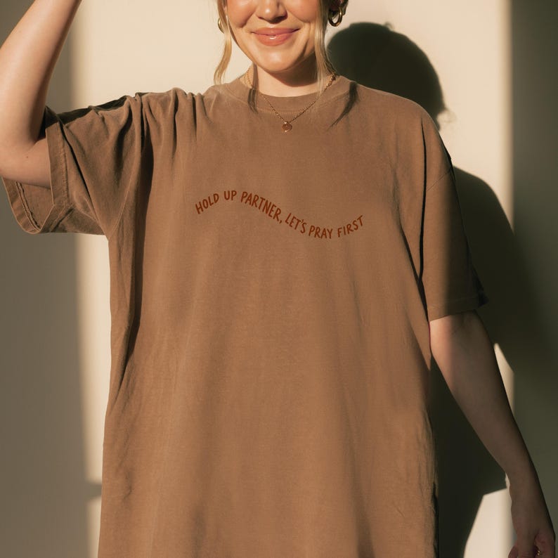 May include: A brown t-shirt with the text "HOLD UP PARTNER, LET'S PRAY FIRST" in a curved, rust-colored font. The shirt has short sleeves and a crew neck. The fabric appears soft and comfortable.