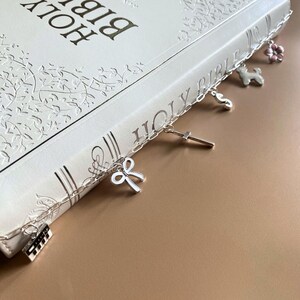 May include: A white Bible with embossed floral patterns, featuring a silver chain bookmark adorned with charms. Charms include a cross, a bow, a flower, a dog, and a piano key design. The book's title, "HOLY BIBLE," is visible.
