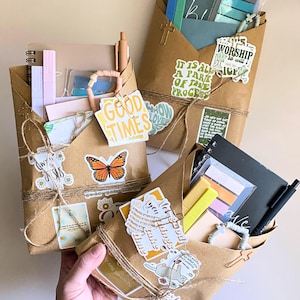 Puede incluir: Tres paquetes de papel marrón atados con hilo, llenos de artículos de papelería, pegatinas y bolígrafos. Cada paquete está decorado con pegatinas que tienen citas e imágenes inspiradoras, incluyendo una mariposa y las palabras "Good Times".