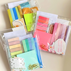 以下が含まれることがあります： 文房具が入った透明なビニール袋。袋には、カラフルな付箋、蛍光ペン、ペーパークリップ、ステッカーが入っています。一部のステッカーには「I WILL GIVE YOU THE GIFT」と「EACH DAY」という文字が書かれています。