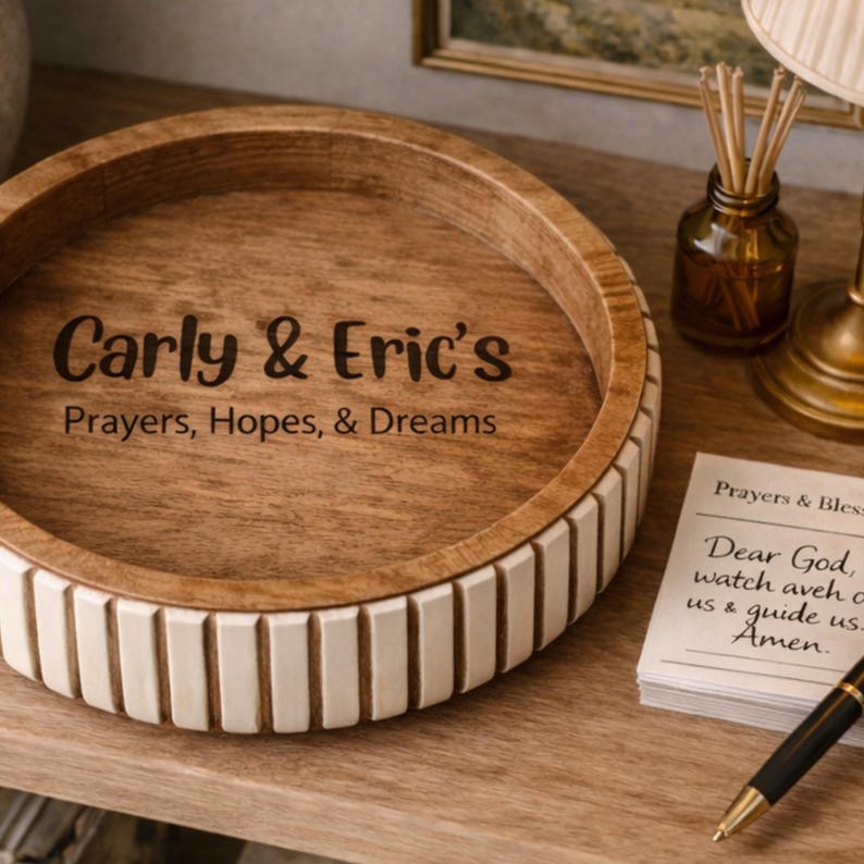 May include: A round wooden tray with the text "Carly & Eric's Prayers, Hopes, & Dreams" engraved on the light-colored wood. The tray has a decorative white and wood-toned rim. A small stack of papers with text and a pen are on the right.