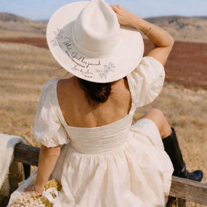 Puede incluir: Un sombrero fedora color crema con ala ancha, con detalles florales y el texto "What God has joined, let no one separate" con una fecha. El sombrero se usa al aire libre.