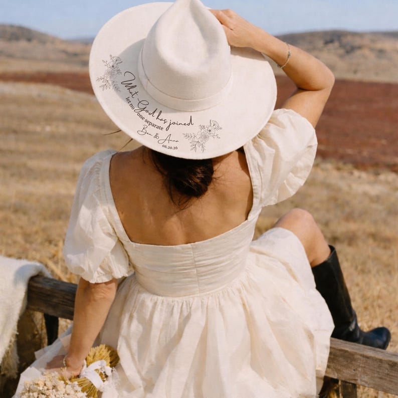 Puede incluir: Un sombrero fedora color crema con ala ancha, con detalles florales y el texto "What God has joined, let no one separate" con una fecha. El sombrero se usa al aire libre.