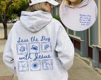 Puede incluir: Una sudadera con capucha gris claro con texto y gráficos en azul. La parte trasera muestra la frase Seas the Day with Jesus sobre seis iconos cuadrados de vida marina. El pecho incluye el texto Mightier than the waves of the sea is His love for you. Psalm 93:4.