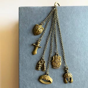 May include: A bronze-toned bookmark with dangling charms. The charms include a shield, sword, helmet, and other medieval-themed elements. The bookmark is attached to a gray surface.