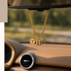 May include: A gold-toned chain hanging from a rearview mirror with three charms: a circular letter T, an open book, and a cross. Below, a rectangular cream-colored case features the text: If the stars are made to worship so will I.