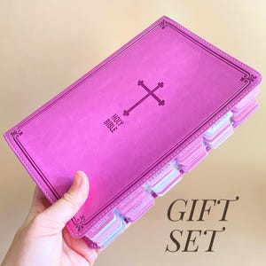 Może przedstawiać: Różowa Biblia z krzyżem i napisem "Holy Bible" na okładce. Książka ma zakładki z etykietami, a słowa "GIFT SET" są wydrukowane w prawym dolnym rogu.