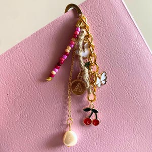 May include: A gold-toned bookmark with a beaded accent and dangling charms. Charms include a gold initial "A" pendant, a white butterfly, red cherries, and a faux pearl. The bookmark is attached to a pink surface.