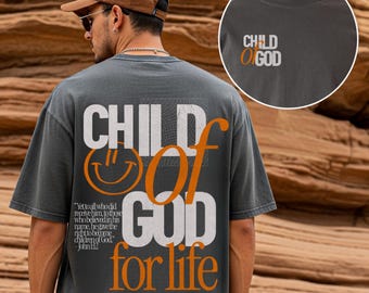 May include: Dark gray t-shirt with the text "CHILD OF GOD for life" in white and orange. The shirt features a smiley face graphic and a Bible verse. The shirt is a casual, oversized fit.
