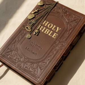 May include: A brown leather Holy Bible with gold lettering and embossed patterns. The cover reads "Holy Bible" and "King James Version". A decorative bronze bookmark with charms hangs from the top. The name "Michael" is embossed on the cover.