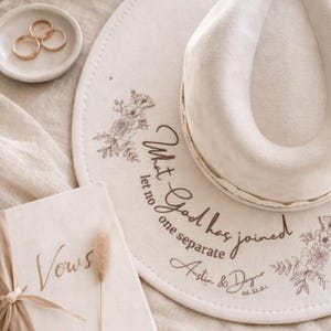 Puede incluir: Un sombrero fedora color crema con detalles florales y la inscripci&oacute;n "What God has joined let no one separate". Un plato peque&ntilde;o contiene tres anillos dorados. Tambi&eacute;n se ve un libro con la palabra "Vows".