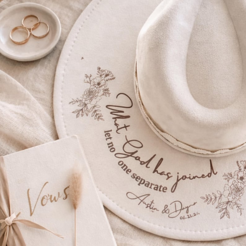 Puede incluir: Un sombrero fedora color crema con detalles florales y la inscripci&oacute;n "What God has joined let no one separate". Un plato peque&ntilde;o contiene tres anillos dorados. Tambi&eacute;n se ve un libro con la palabra "Vows".