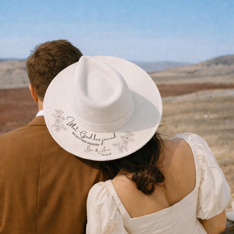 Puede incluir: Un sombrero fedora blanco con ala ancha. El sombrero presenta dise&ntilde;os florales y el texto "What God has joined, let no one separate", junto con nombres y una fecha. Un accesorio de boda.
