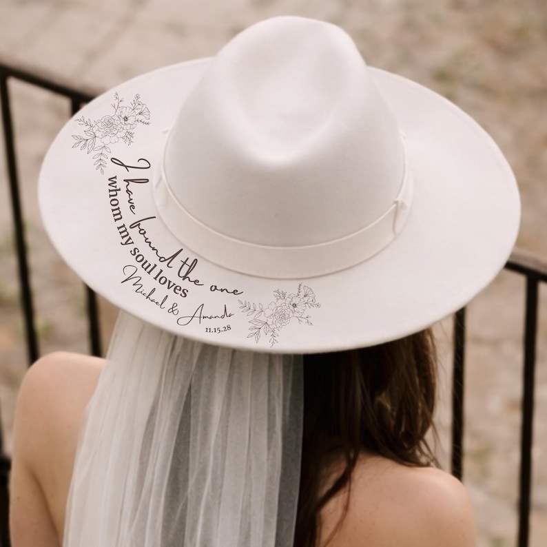 Puede incluir: Un sombrero fedora blanco con un dise&ntilde;o floral y el texto "I have found the one whom my soul loves Michael & Amanda 11.15.28". El sombrero tiene una cinta blanca. Una persona lleva el sombrero con un velo.