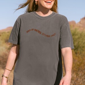 May include: Dark gray t-shirt with the text "HOLD UP PARTNER, LET'S PRAY FIRST" in a curved, rust-colored font. The shirt has short sleeves and a relaxed fit. The background shows a blurred outdoor scene with mountains.