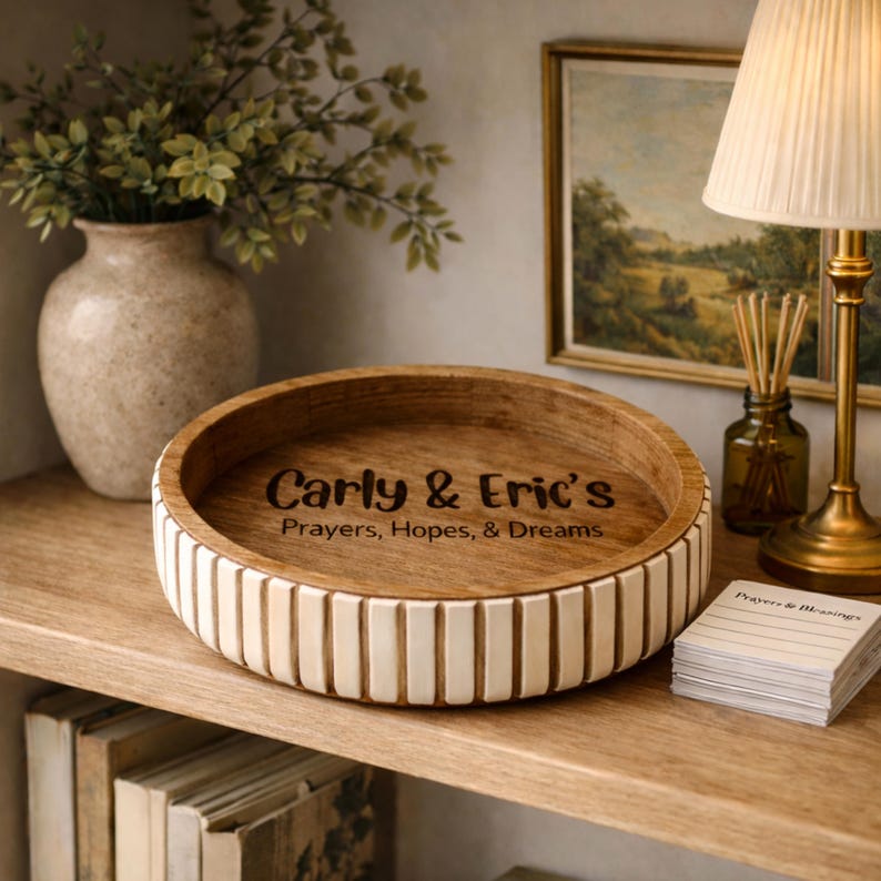 May include: A round wooden decorative bowl with a light brown interior and a white and brown striped exterior. The bowl has the text "Carly & Eric's Prayers, Hopes, & Dreams" engraved on the inside. The bowl is on a wooden shelf.