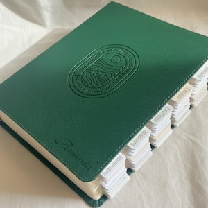 Puede incluir: Un libro verde encuadernado en cuero con el texto en relieve "THIS HEAVENS DECLARE THE GLORY OF GOD". El libro tiene pestañas etiquetadas y el nombre "Amanda" está grabado en la portada.