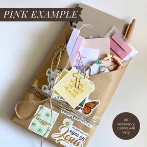 May include: A brown paper package tied with twine containing a spiral notebook, pens, and stickers. The stickers feature butterflies, daisies, and religious text including "By His wounds we are healed" and "We must keep our eyes on Jesus."