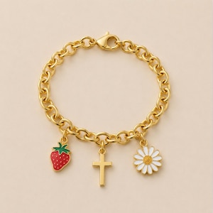 May include: A gold-toned chain link bracelet featuring three dangling charms: a red and green enameled strawberry, a plain gold-toned cross, and a white and yellow enameled daisy flower. The bracelet is secured with a lobster claw clasp.