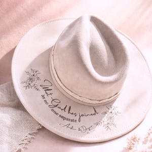 Puede incluir: Sombrero fedora beige claro con ala ancha. El sombrero presenta un dise&ntilde;o floral y el texto "What God has joined let no one separate" con nombres y una fecha. El sombrero es de color neutro.
