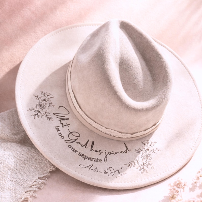 Puede incluir: Sombrero fedora beige claro con ala ancha. El sombrero presenta un dise&ntilde;o floral y el texto "What God has joined let no one separate" con nombres y una fecha. El sombrero es de color neutro.