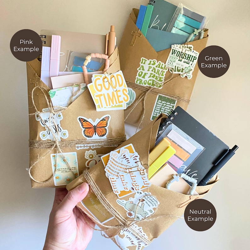 May include: Three brown paper gift envelopes tied with twine, each containing stationery items like notebooks, pens, and sticky notes. The envelopes are decorated with various stickers featuring text like GOOD TIMES and butterflies, plus small cross charms.