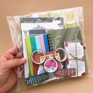 May include: A clear plastic bag containing a stationery set with a SOAP METHOD notebook, five colorful markers in lime, teal, blue, pink, and dark blue, three rolls of patterned washi tape, a small spiral notebook, and a card with religious text.