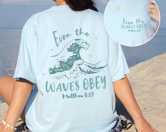 May include: Pale blue t-shirt featuring the text "Even the Waves Obey" and a wave graphic. The text "Matthew 8:27" is below the wave design. The shirt has a loose fit and short sleeves.