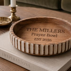 Puede incluir: Un cuenco redondo de madera con interior marrón natural y exterior pintado de blanco con nervaduras verticales. El interior está grabado con el texto THE MILLERS Prayer Bowl EST 2026. El cuenco tiene un diseño circular poco profundo.