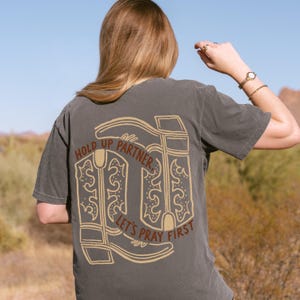 May include: Dark gray t-shirt with a tan graphic of cowboy boots and the text "HOLD UP PARTNER, LET'S PRAY FIRST." The shirt is short-sleeved and has a relaxed fit. The background shows a desert landscape.