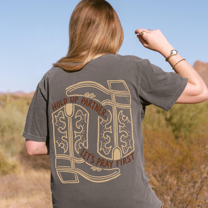 May include: Dark gray t-shirt with a tan graphic of cowboy boots and the text "HOLD UP PARTNER, LET'S PRAY FIRST." The shirt is short-sleeved and has a relaxed fit. The background shows a desert landscape.