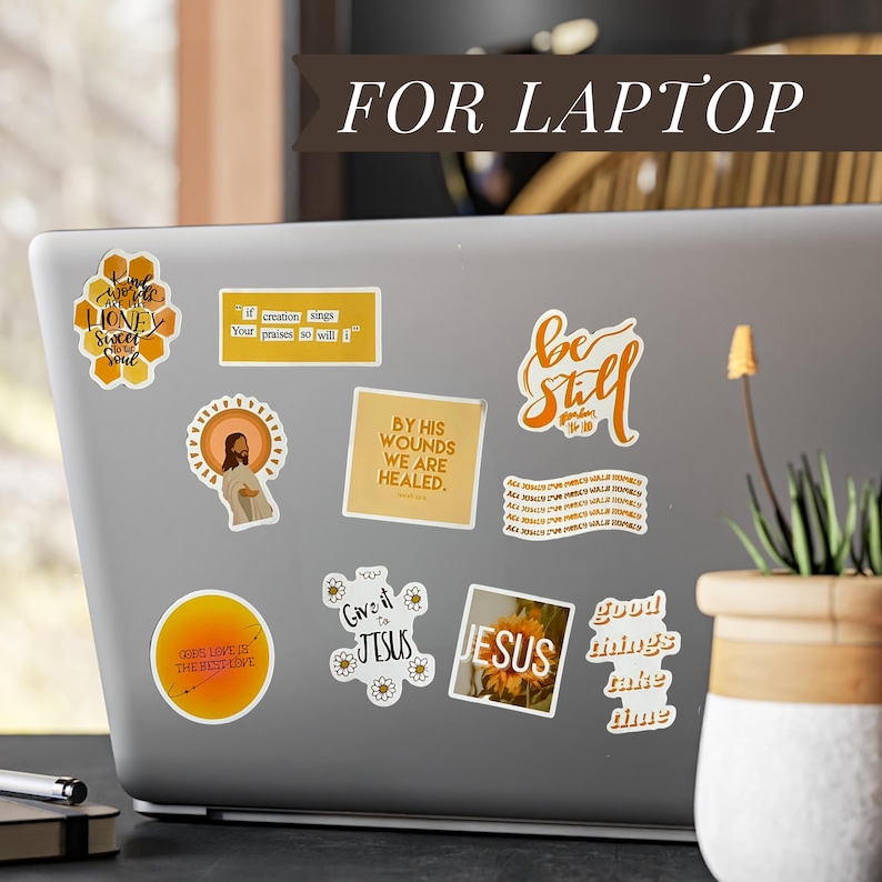 May include: A laptop covered in various stickers with religious themes. The stickers feature phrases like "Be Still", "Jesus", and "By His Wounds We Are Healed". Other stickers have floral and honeycomb designs.