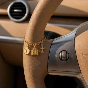 May include: A gold-toned chain bracelet featuring three dangling charms: a circular pendant with the letter T, a small open book, and a simple cross. The bracelet is constructed with round links and has a polished metallic finish throughout.