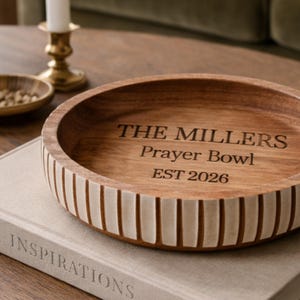 May include: A wooden prayer bowl with the text "THE MILLERS Prayer Bowl EST 2026" engraved on the inside. The bowl has a light brown interior and a darker brown exterior with vertical white stripes. The bowl is sitting on a book.