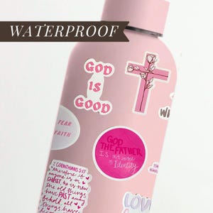 May include: A pink, waterproof water bottle covered in stickers. The stickers feature phrases like "God is Good," a pink cross with floral accents, and other inspirational quotes. The bottle is designed for carrying water or other beverages.