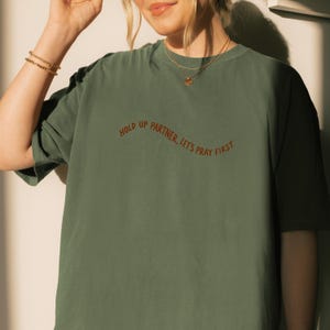May include: Olive green t-shirt with the text "HOLD UP PARTNER, LET'S PRAY FIRST" in a curved, rust-colored font. The shirt has a relaxed fit and short sleeves. The image shows the upper body of a person wearing the shirt.