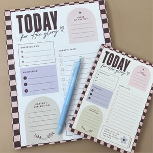 May include: Two daily planner sheets with a brown and white checkered border, featuring the text "TODAY for His glory" and sections for "Grateful For," "Today's Plan," "Priorities," and "Prayer + Reflection," accompanied by a light blue pen.