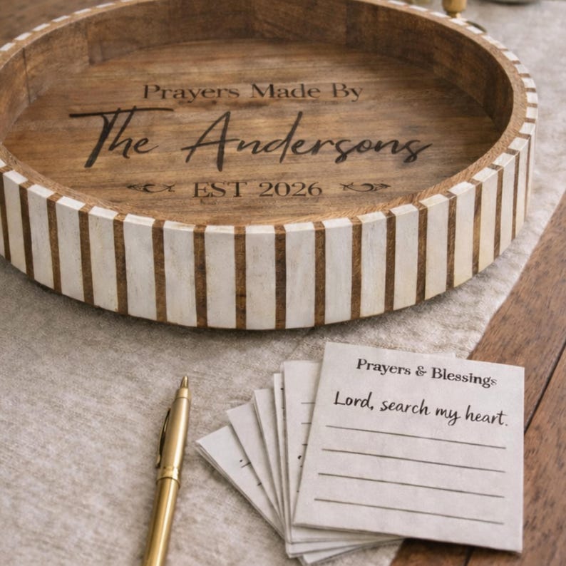 May include: A round wooden tray with a light brown interior and a white and brown striped exterior. The tray has the text "Prayers Made By The Andersons EST 2026" engraved on the inside. A gold pen and a stack of notecards are also present.