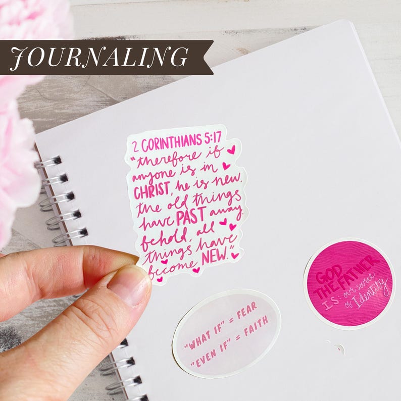 May include: A hand holding a sticker with the text "2 Corinthians 5:17" in pink script, with other stickers on a white notebook. The word "JOURNALING" is in a banner at the top.