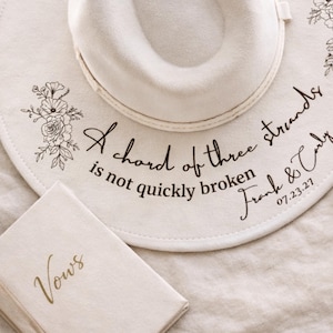 Könnte beinhalten: Cremefarbener Fedora-Hut mit floralen Akzenten und der Aufschrift "A chord of three strands is not quickly broken Frank & Carly 07.23.27." Ein kleines Buch mit dem Wort "Vows" in Gold. Hochzeitsthema.