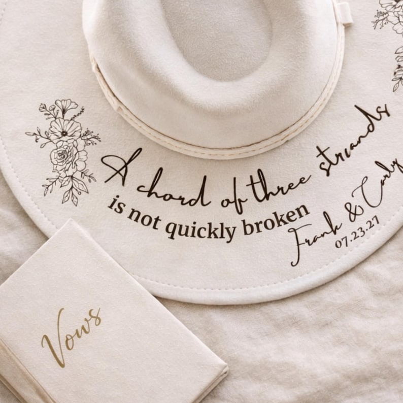 Puede incluir: Sombrero fedora blanco roto con detalles florales y la inscripci&oacute;n "A chord of three strands is not quickly broken Frank & Carly 07.23.27." Un peque&ntilde;o libro con la palabra "Vows" en letras doradas. Tema de boda.