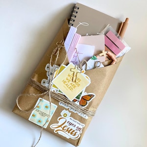 Puede incluir: Un paquete de papel marrón atado con hilo, lleno de artículos de papelería. Incluye un cuaderno de espiral, pegatinas, bolígrafos y pequeñas cruces de madera. Las pegatinas dicen "This wounds we are healed" y "gods got this."