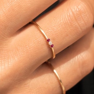 May include: A thin gold ring featuring a central clear stone flanked by two small red stones. The band is smooth and metallic, worn on a finger alongside a second plain, thin gold band. The design is delicate, showcasing a minimalist arrangement of three small gemstones set horizontally on the slender gold ring band.