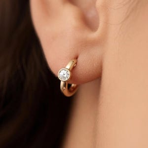 May include: A small gold hoop earring featuring a single round, bezel-set clear gemstone. The earring is secured through the earlobe, showcasing a polished metallic finish and a minimalist circular design that sits flush against the skin.