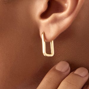 May include: A close-up of a gold-colored, rectangular hoop earring. The earring is worn on the ear of a person with tan skin. The earring is a simple, modern design. A hand is touching the ear.